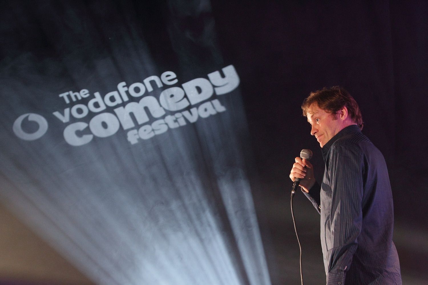 Ardal O'Hanlon onstage at the Vodafone Comedy Festival, with the Festival logo projected onto the wall in front of him