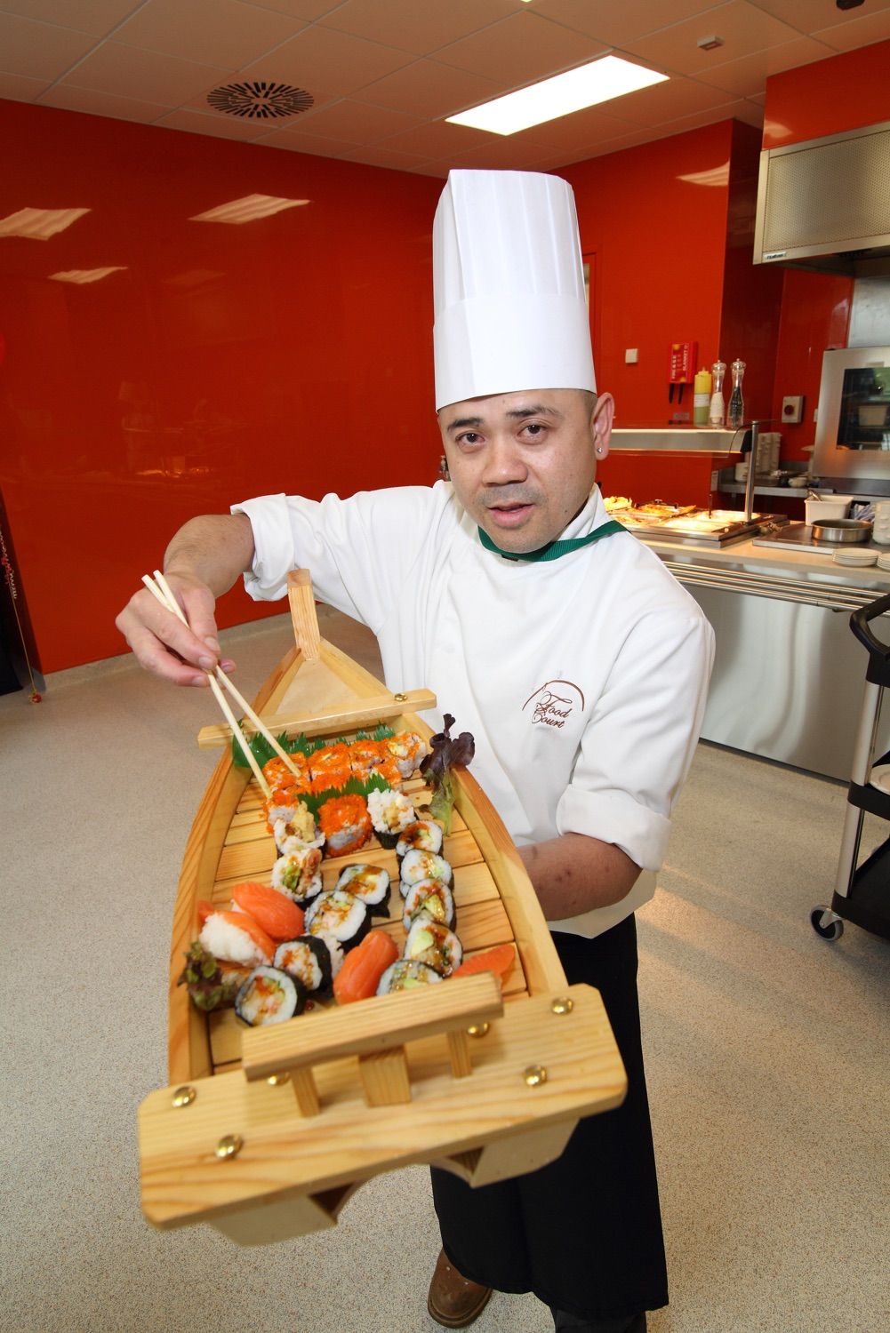 A chef at a company event uses chop sticks to offer a taste of freshly prepared sushi fro a wooden boat platter