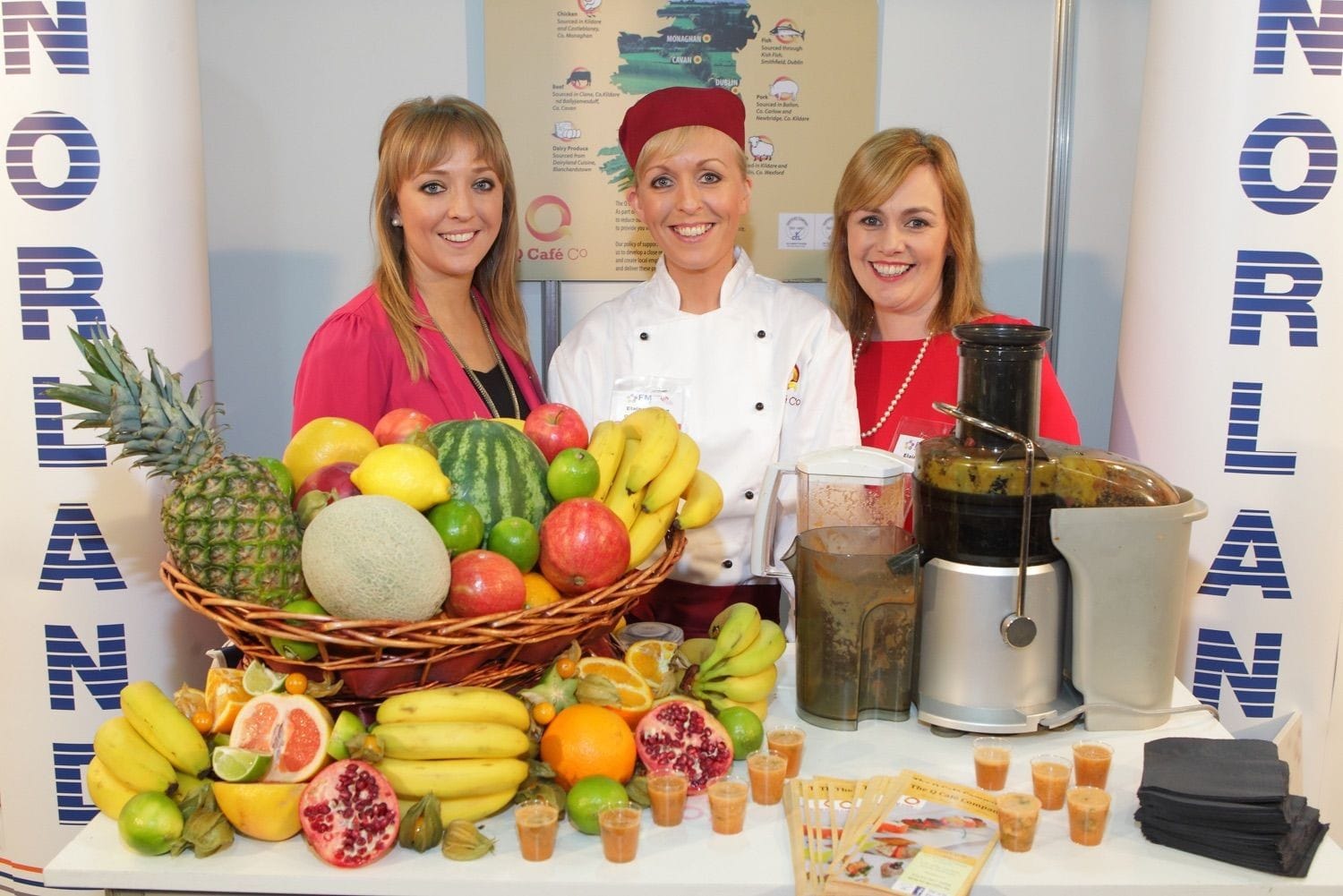 Three lovely ladies stand behind a stunning display of colourful fruit to promote fuit smoothies