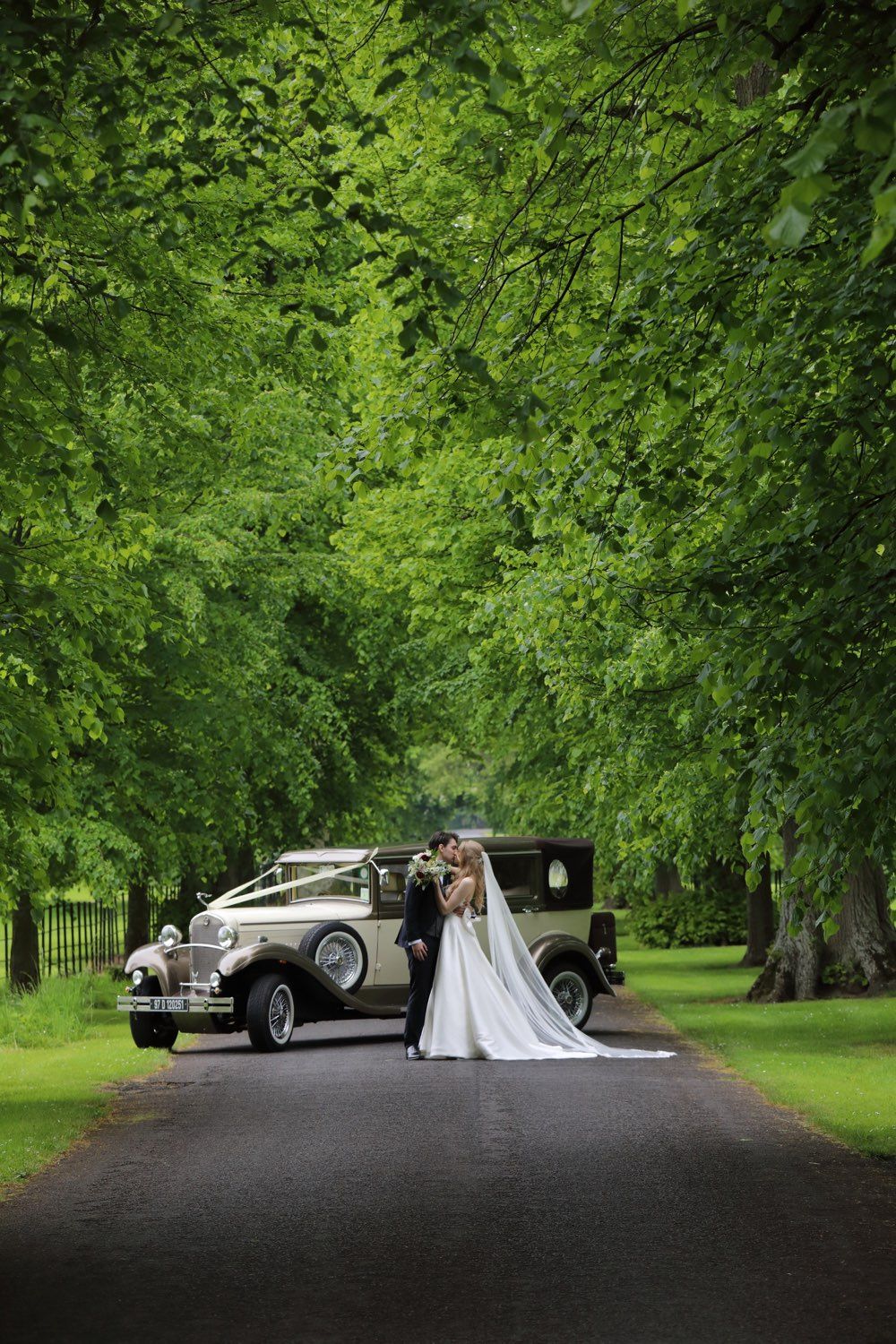 The bride and groom standing with the car behind them on the tree lined avenue of Luttrelstown Castel