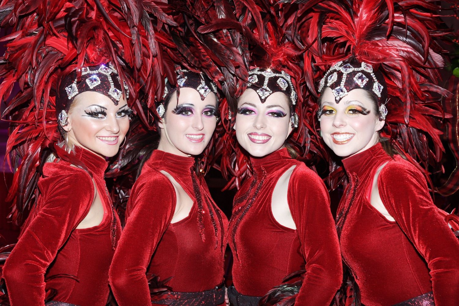 A close up photograph of four young ladies dressed in red velvet leotards and feathers smile for the camera